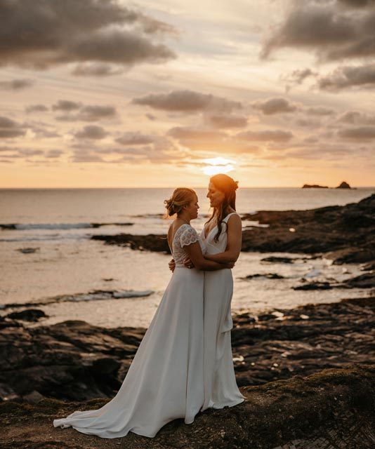Emma and Annie embracing during their wedding in front of the coast and a sunset.